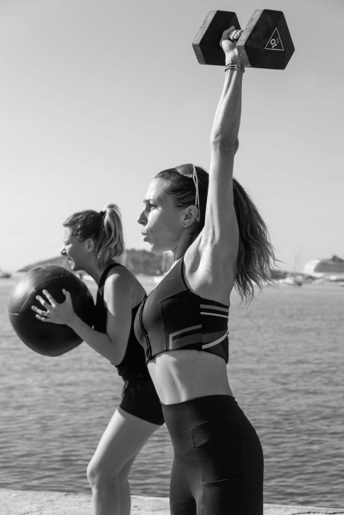 Two women doing an exercice workout in the beach. One doing with a dumbell, the other one with a ball.